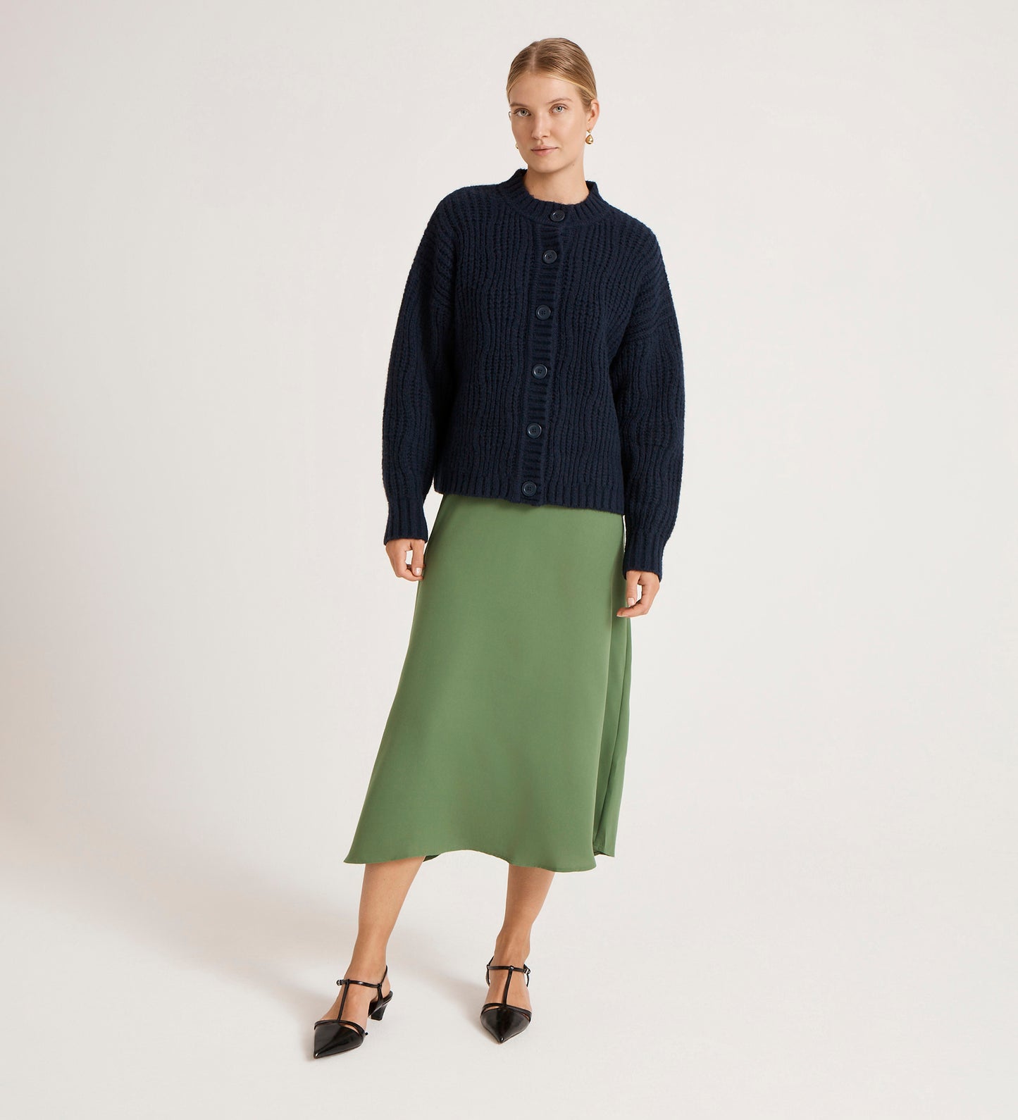 A woman stands against a plain background, wearing the Ailsa Navy Knitted Cardigan, paired with a light green midi skirt and black pointed-toe heels with ankle straps.