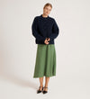 A woman stands against a plain background, wearing the Ailsa Navy Knitted Cardigan over a long green skirt and black pointed-toe heels. She faces the camera with her hands clasped in front of her.