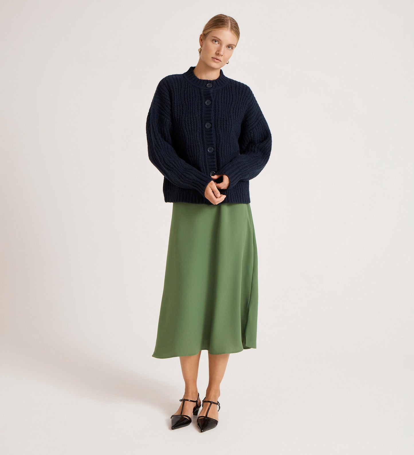 A woman stands against a plain background, wearing the Ailsa Navy Knitted Cardigan over a long green skirt and black pointed-toe heels. She faces the camera with her hands clasped in front of her.