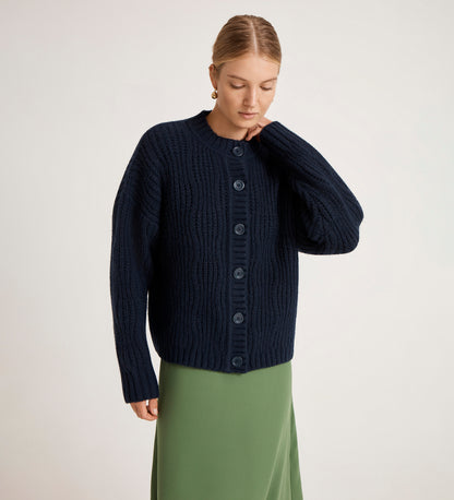 Wearing the Ailsa Navy Knitted Cardigan and a long, light green skirt, a woman stands against a plain light background, gazing down with one hand near her neck.