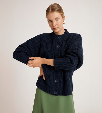 A blonde-haired woman wears the Ailsa Navy Knitted Cardigan and a green skirt, standing against a plain light background with one arm folded over the other.