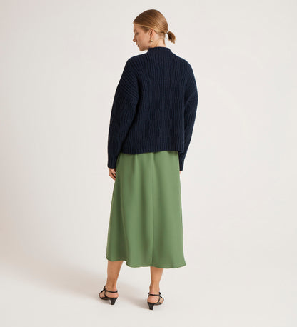 A person faces away, wearing the Ailsa Navy Knitted Cardigan with a green midi skirt and black heeled sandals. Their hair is tied back, set against a plain light background.