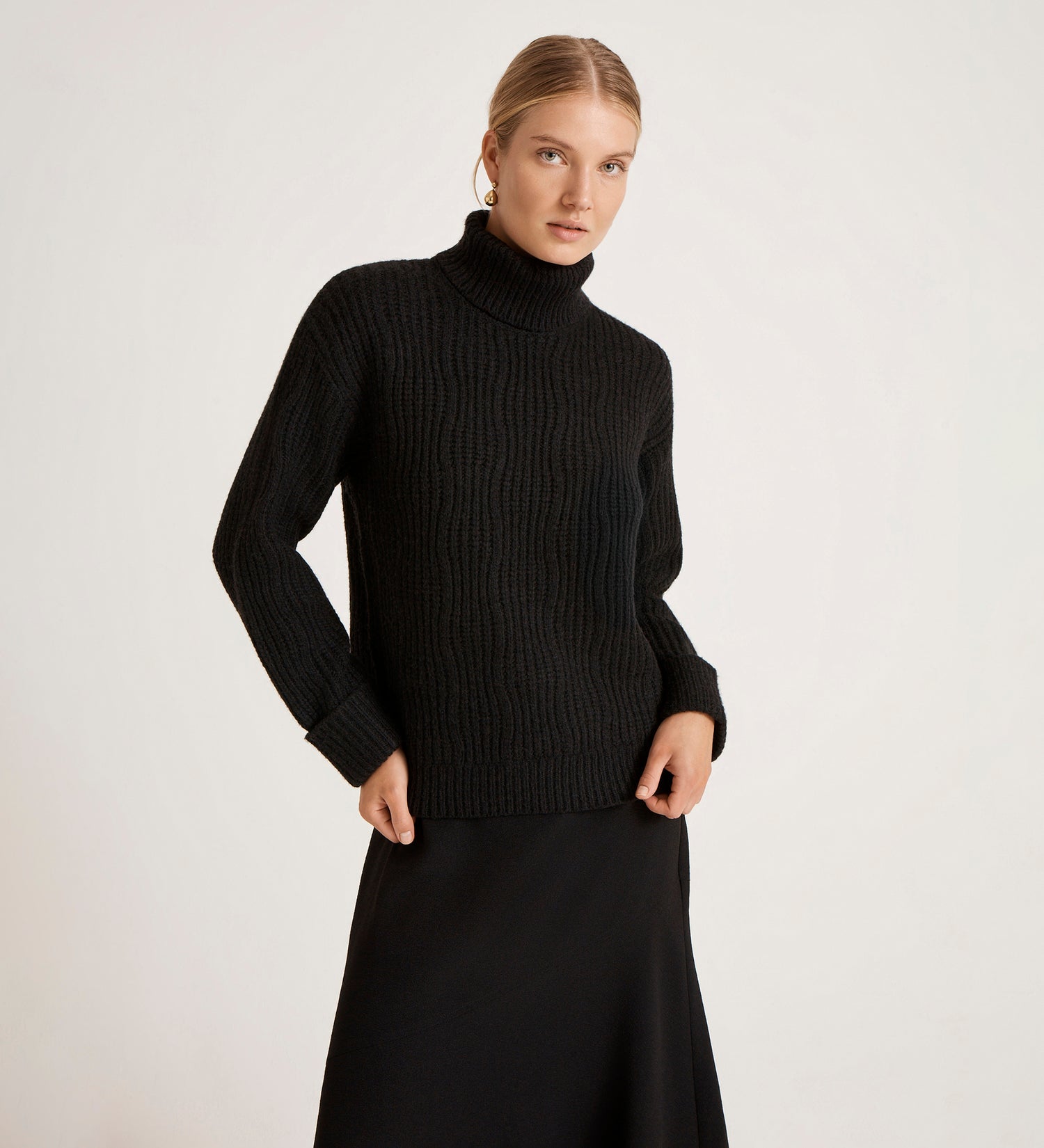 A woman with blonde hair pulled back stands against a plain background, wearing the Albie Black Roll Neck Jumper and a long black skirt. She looks slightly to the side with a neutral expression.
