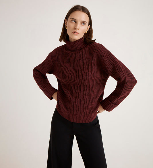 A woman with straight brown hair wears the Albie Burgundy Roll Neck Jumper and black pants, standing with her hands on her hips against a plain light background.