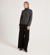 A person stands against a plain background wearing the Albie Charcoal Roll Neck Jumper, black wide-leg pants, and dark pointed shoes. Their blonde hair is pulled back as they look slightly to the side.