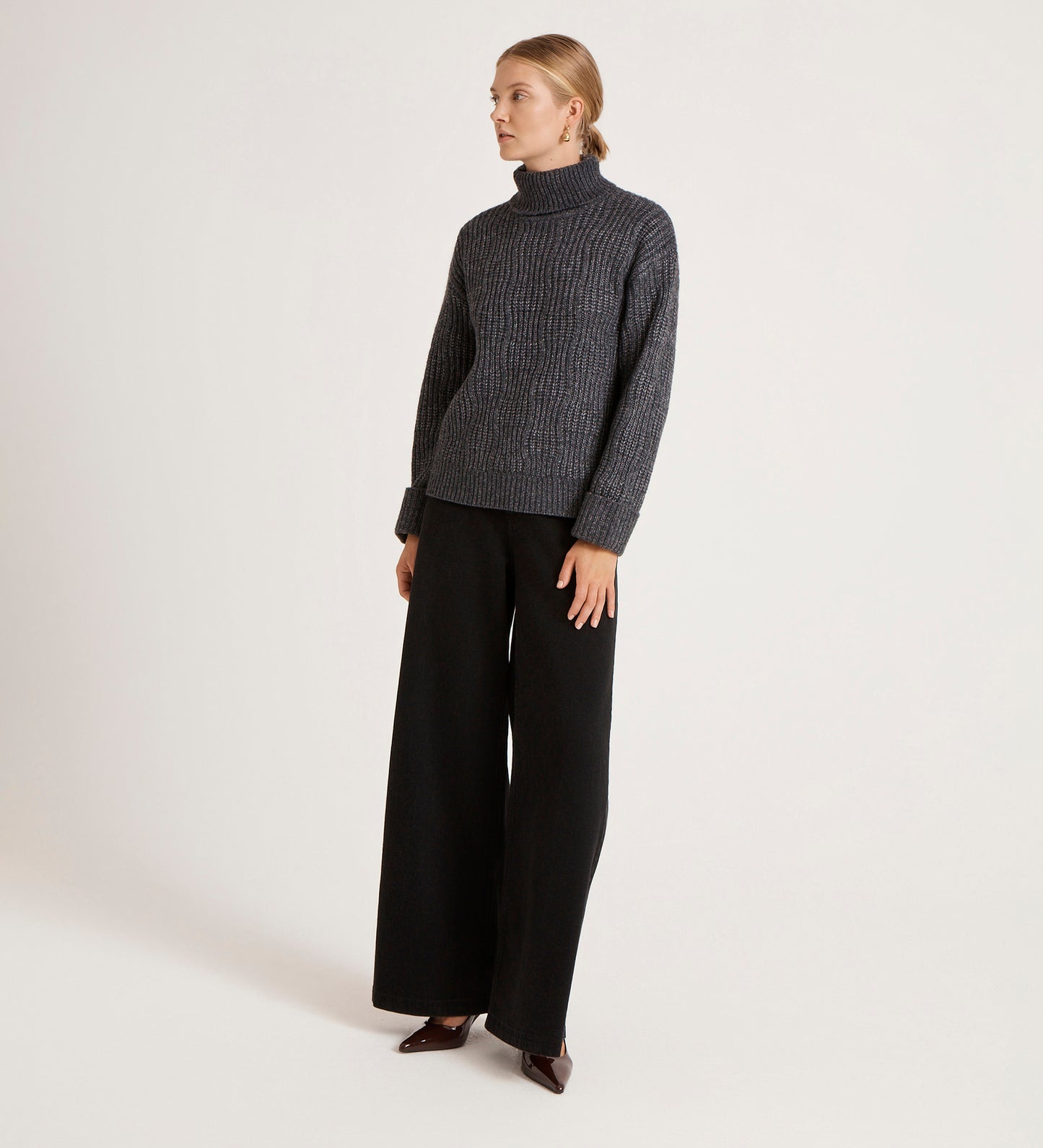 A person stands against a plain background wearing the Albie Charcoal Roll Neck Jumper, black wide-leg pants, and dark pointed shoes. Their blonde hair is pulled back as they look slightly to the side.