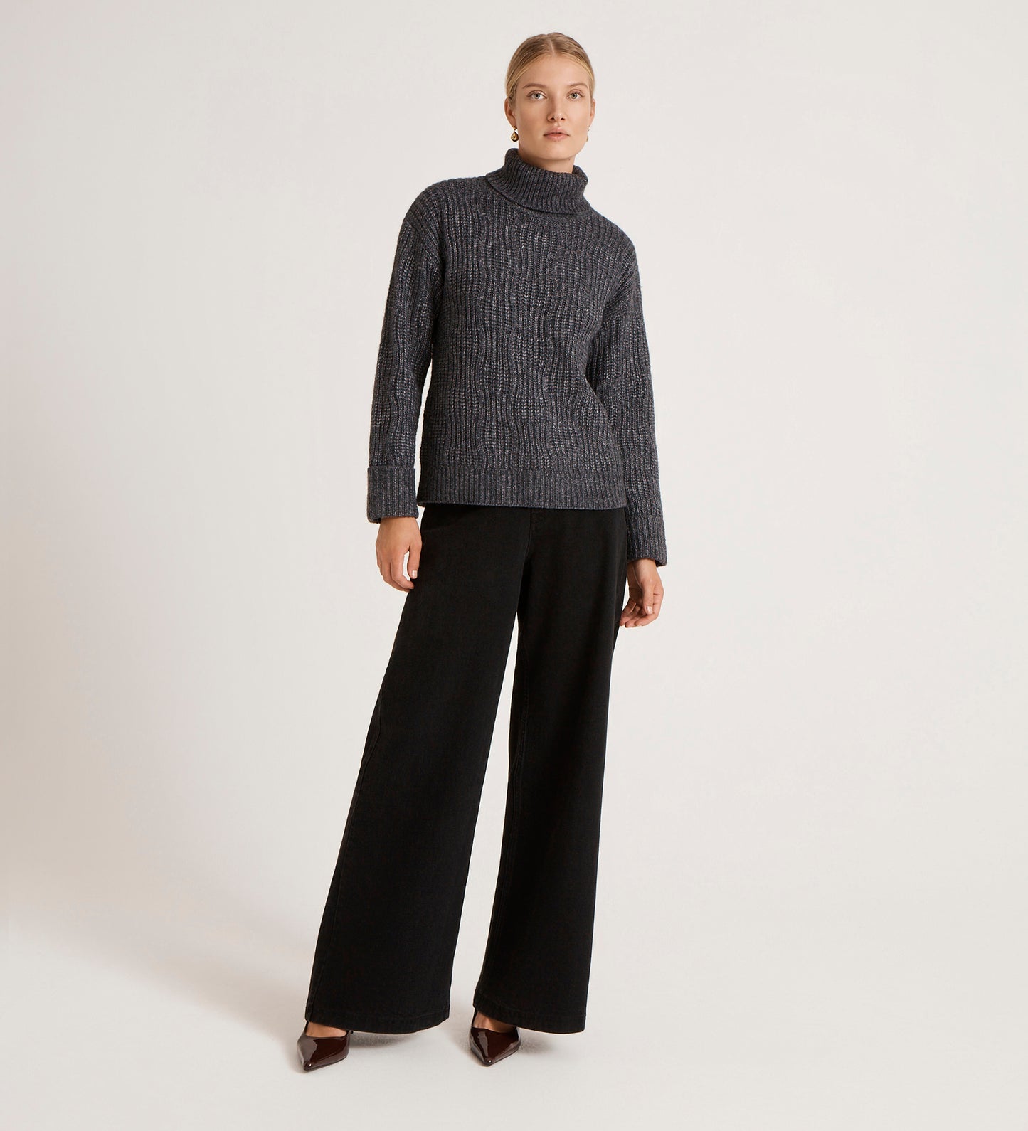 A woman with blonde hair pulled back stands against a plain background, wearing the Albie Charcoal Roll Neck Jumper and wide-leg black pants with brown pointed shoes, her expression neutral.
