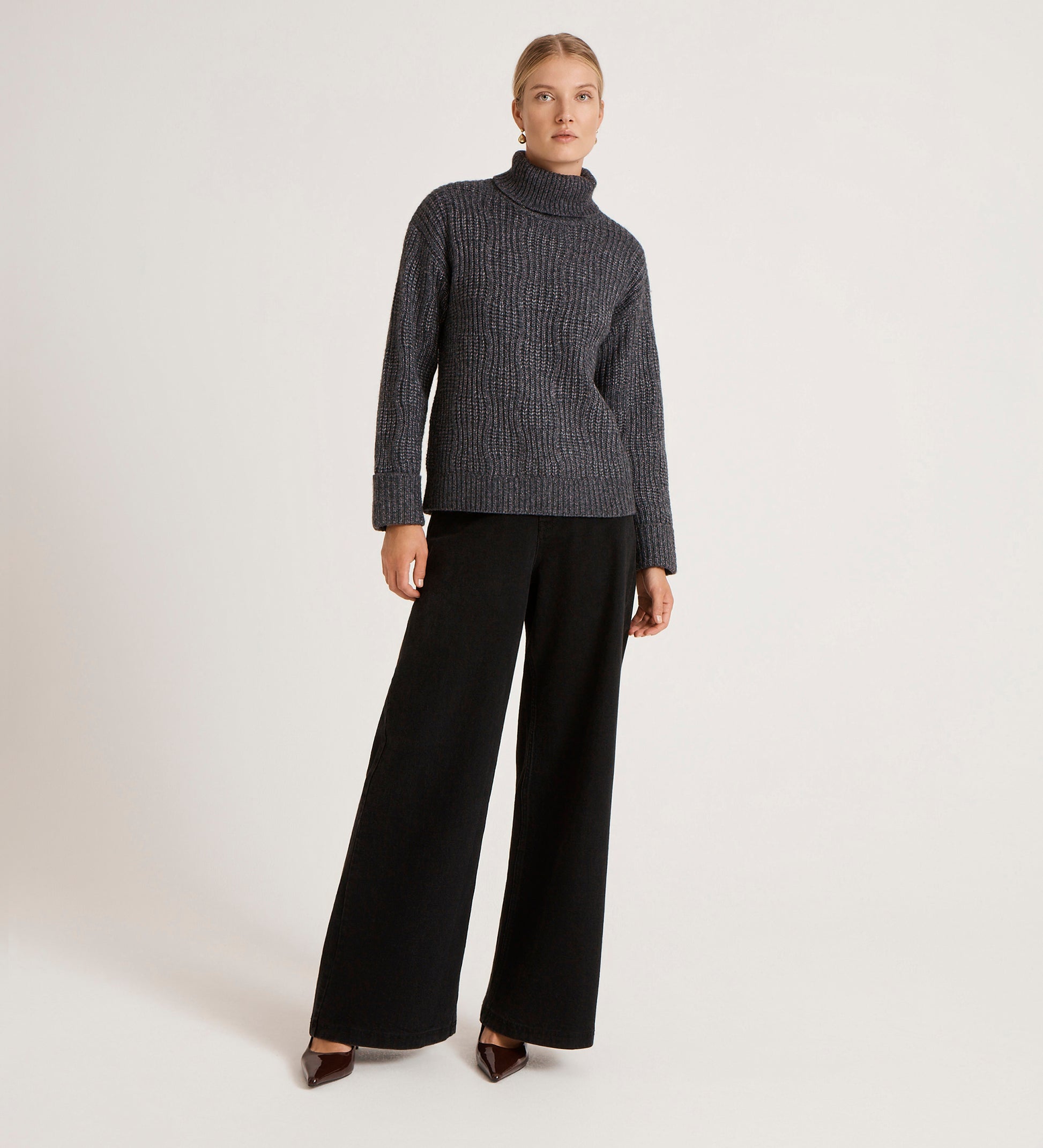 A woman with blonde hair pulled back stands against a plain background, wearing the Albie Charcoal Roll Neck Jumper and wide-leg black pants with brown pointed shoes, her expression neutral.