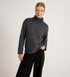 A woman poses against a plain white background wearing the Albie Charcoal Roll Neck Jumper with a dark gray ribbed neck and loose black pants, one hand in her pocket.