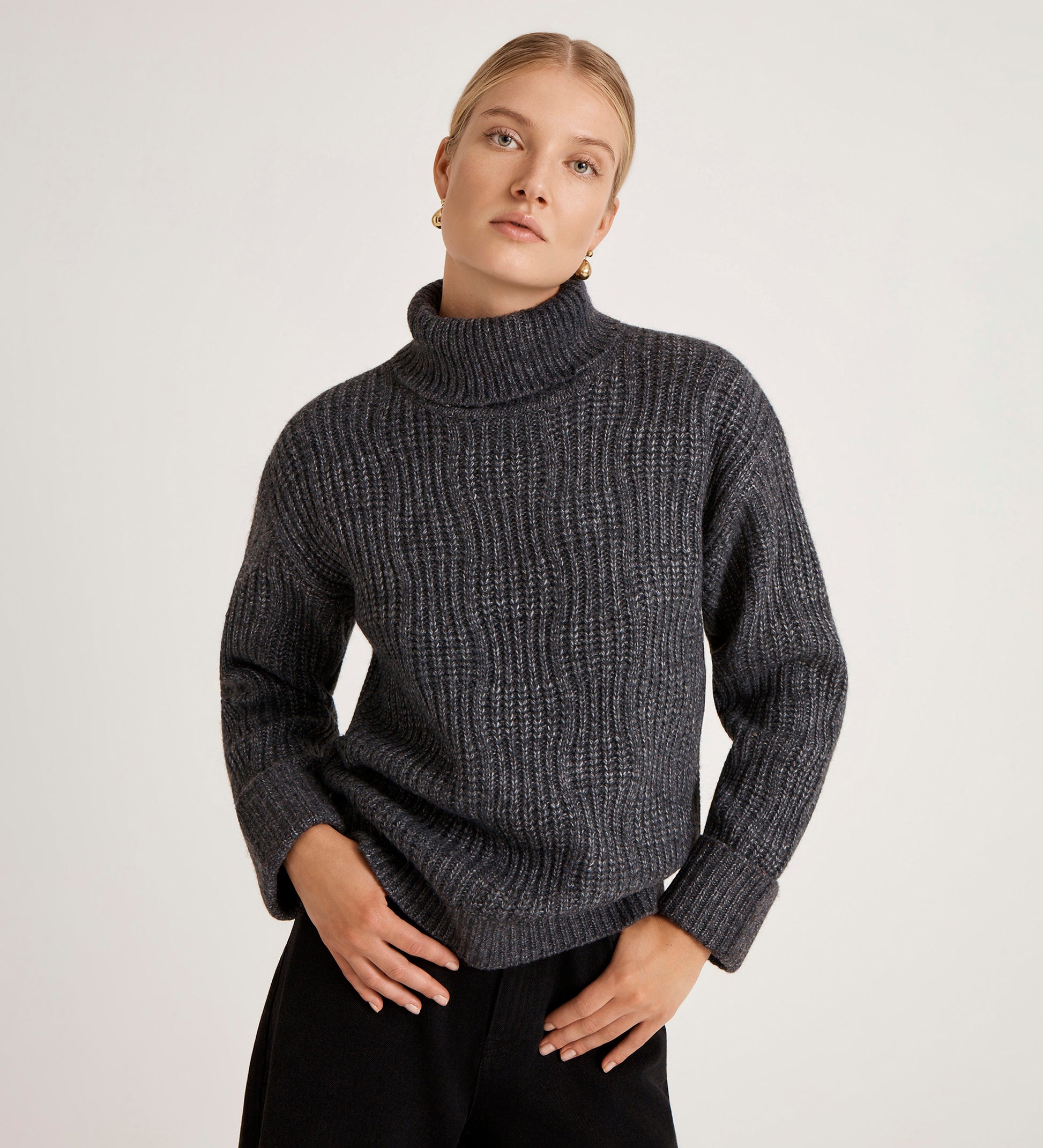 A woman with blonde hair in a low bun wears the Albie Charcoal Roll Neck Jumper and black pants, standing against a plain light background with her hands on her hips.