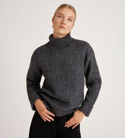 A woman with blonde hair in a low bun wears the Albie Charcoal Roll Neck Jumper and black pants, standing against a plain light background with her hands on her hips.