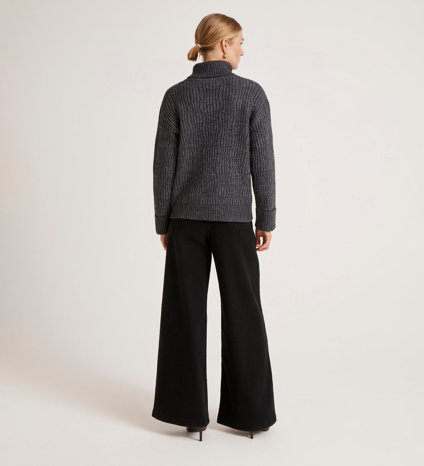 A woman faces away in the Albie Charcoal Roll Neck Jumper, styled with wide-leg black pants and black heels. Her hair is tied in a low bun against a plain white background.