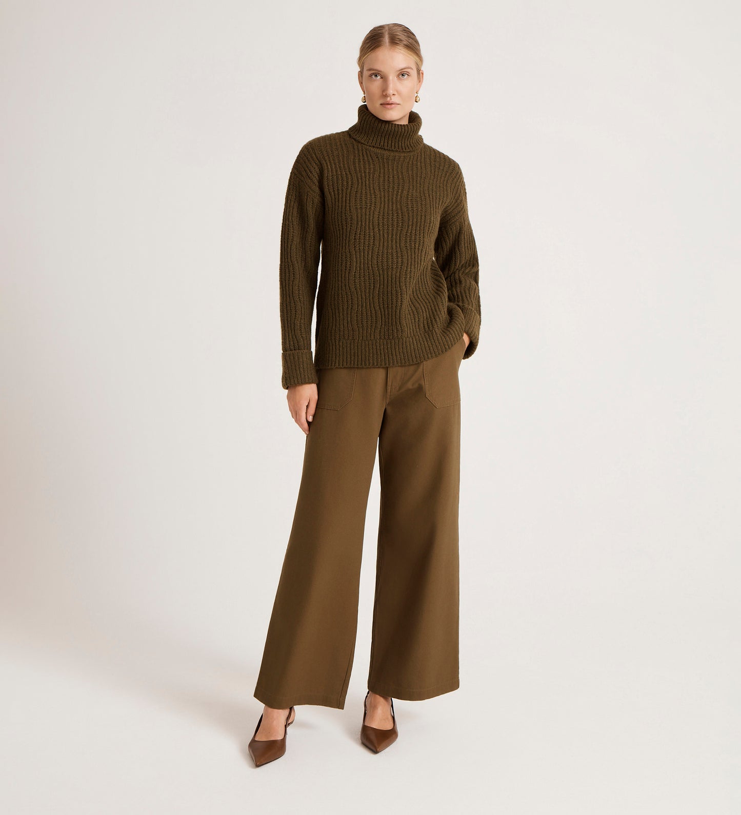 A person stands against a plain background wearing the Albie Khaki Roll Neck Jumper, wide-leg brown pants, and brown heels, with one hand in a pocket and a neutral expression.
