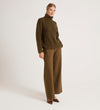 A person stands against a plain background wearing the Albie Khaki Roll Neck Jumper, wide-leg olive trousers, and brown pointed shoes, hands in pockets and a neutral expression.