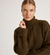 A woman with blonde hair tied back wears the Albie Khaki Roll Neck Jumper and gold earrings. She faces the camera with a neutral expression against a plain light background.