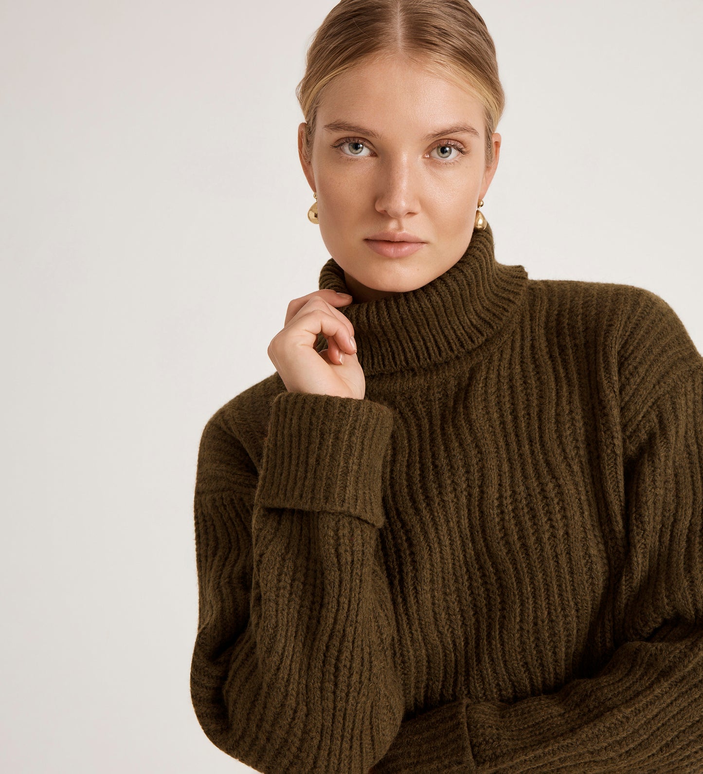 A woman with blonde hair tied back wears the Albie Khaki Roll Neck Jumper and gold earrings. She faces the camera with a neutral expression against a plain light background.