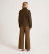 A woman faces away against a plain light background, wearing the Albie Khaki Roll Neck Jumper and wide-leg brown trousers paired with heeled shoes.