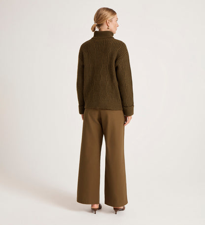 A woman faces away against a plain light background, wearing the Albie Khaki Roll Neck Jumper and wide-leg brown trousers paired with heeled shoes.