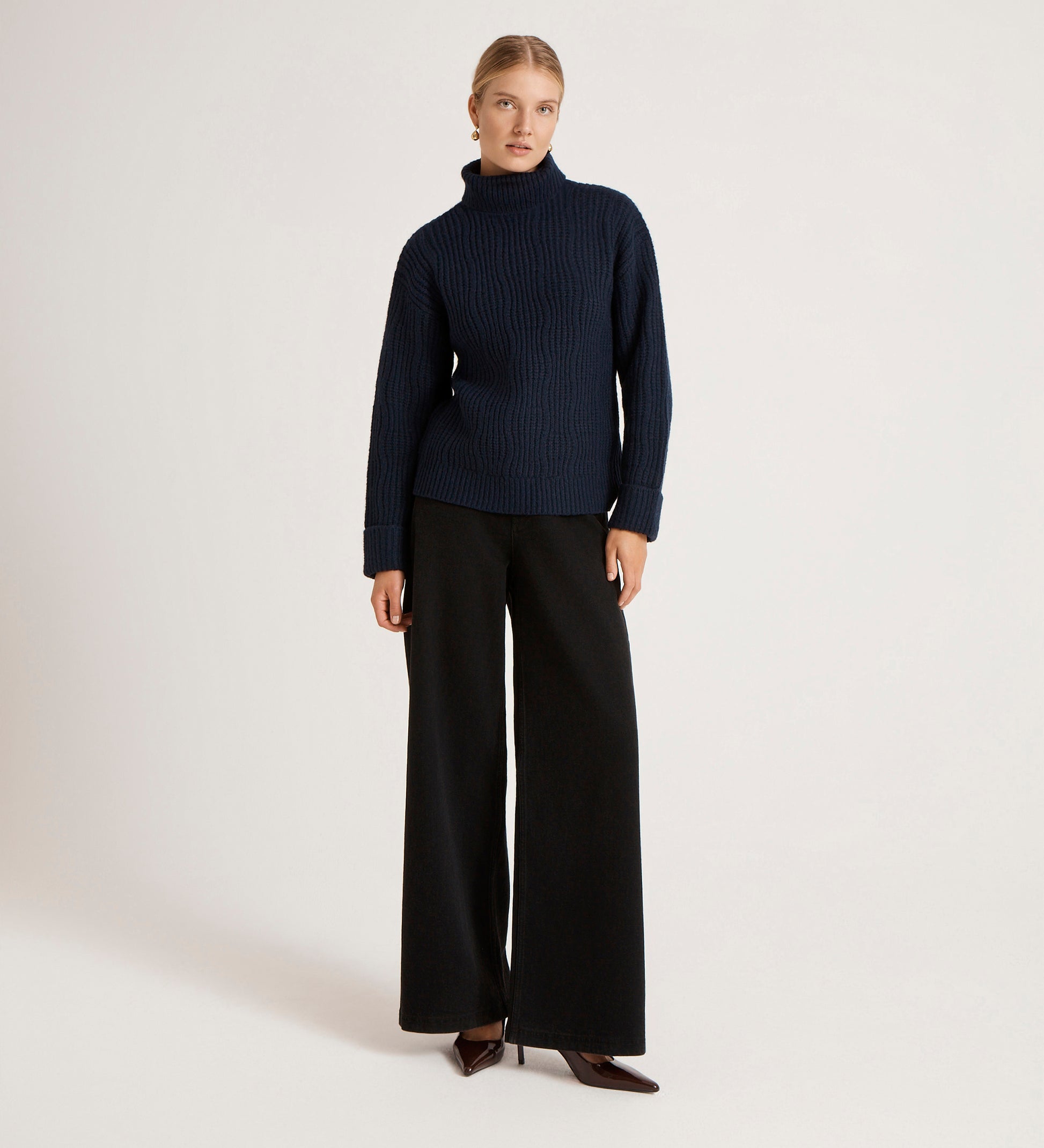 A woman stands against a plain background wearing the Albie Navy Roll Neck Jumper with black wide-leg trousers and brown pointed-toe heels. Her hair is pulled back and her hands rest by her sides.