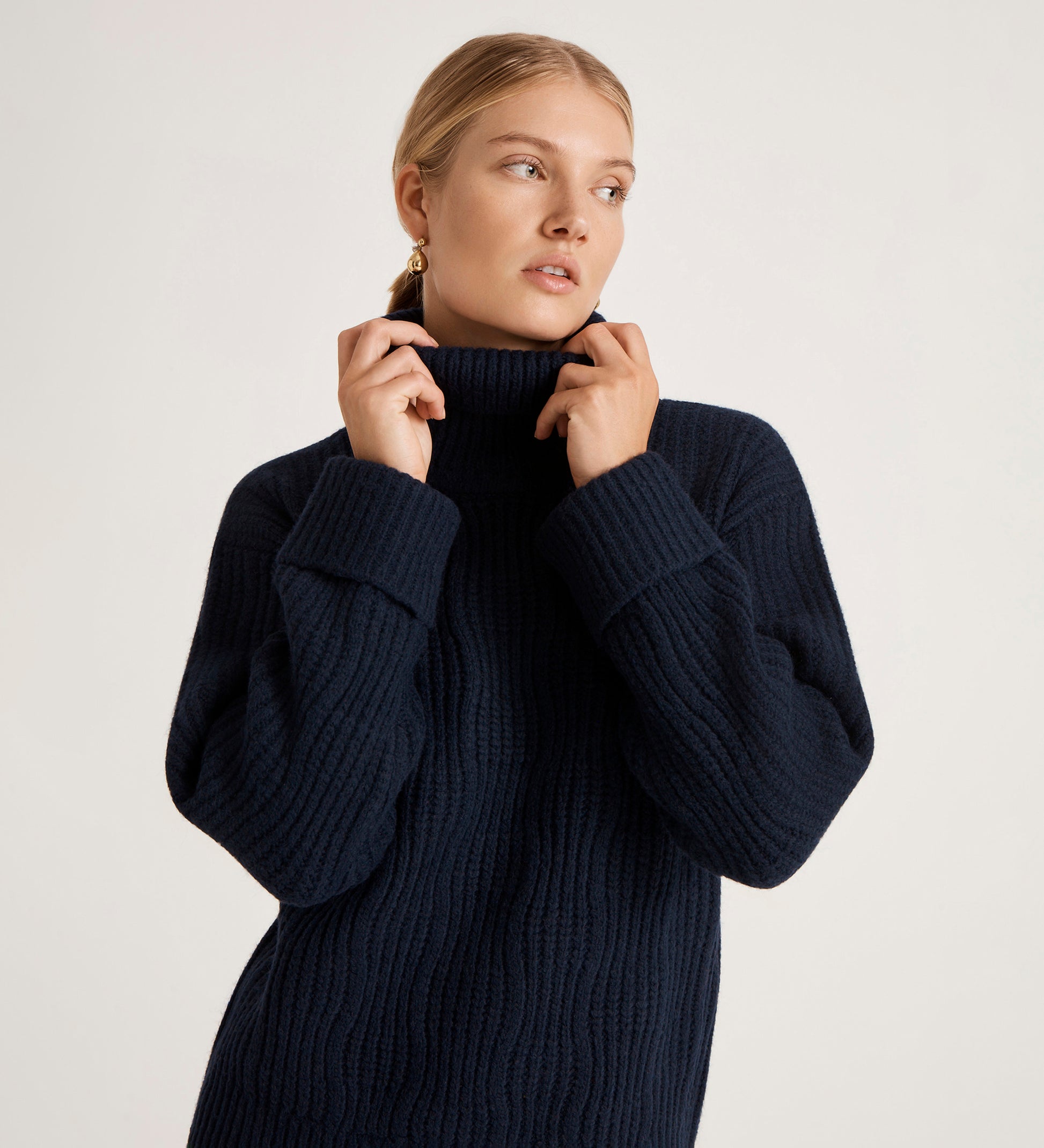 A woman with blonde hair in a low bun wears the Albie Navy Roll Neck Jumper, made from a sustainable yarn blend, holding the cosy collar and looking thoughtfully to the side against a plain light background.