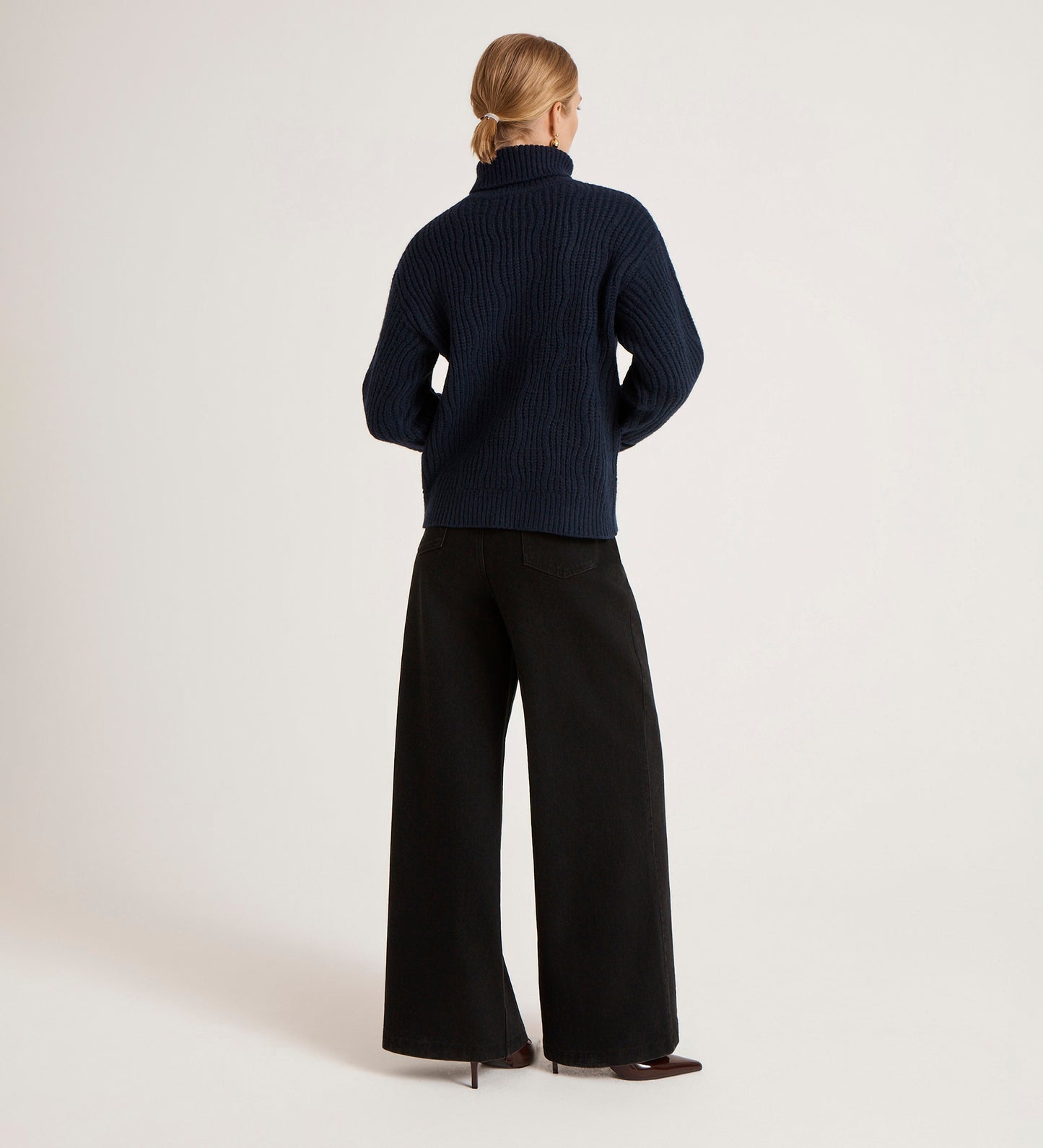 A person stands with their back to the camera, wearing the Albie Navy Roll Neck Jumper styled with wide-leg black pants and a low bun, set against a simple light background.