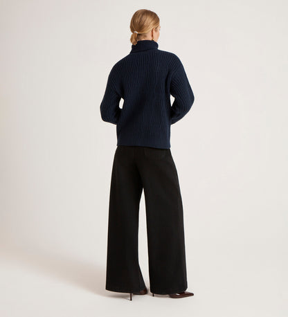 A person stands with their back to the camera, wearing the Albie Navy Roll Neck Jumper styled with wide-leg black pants and a low bun, set against a simple light background.