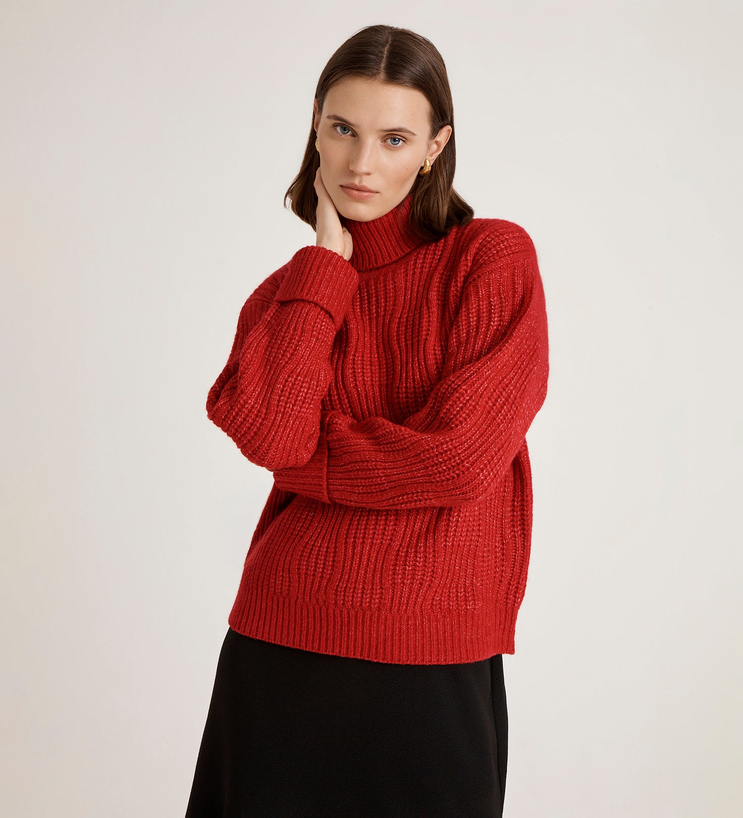 A woman with straight brown hair wears the Albie Red Roll Neck Jumper—a cosy, chunky ribbed sweater—paired with a black skirt. She stands against a plain light background, resting her hand on her chin and looking at the camera.