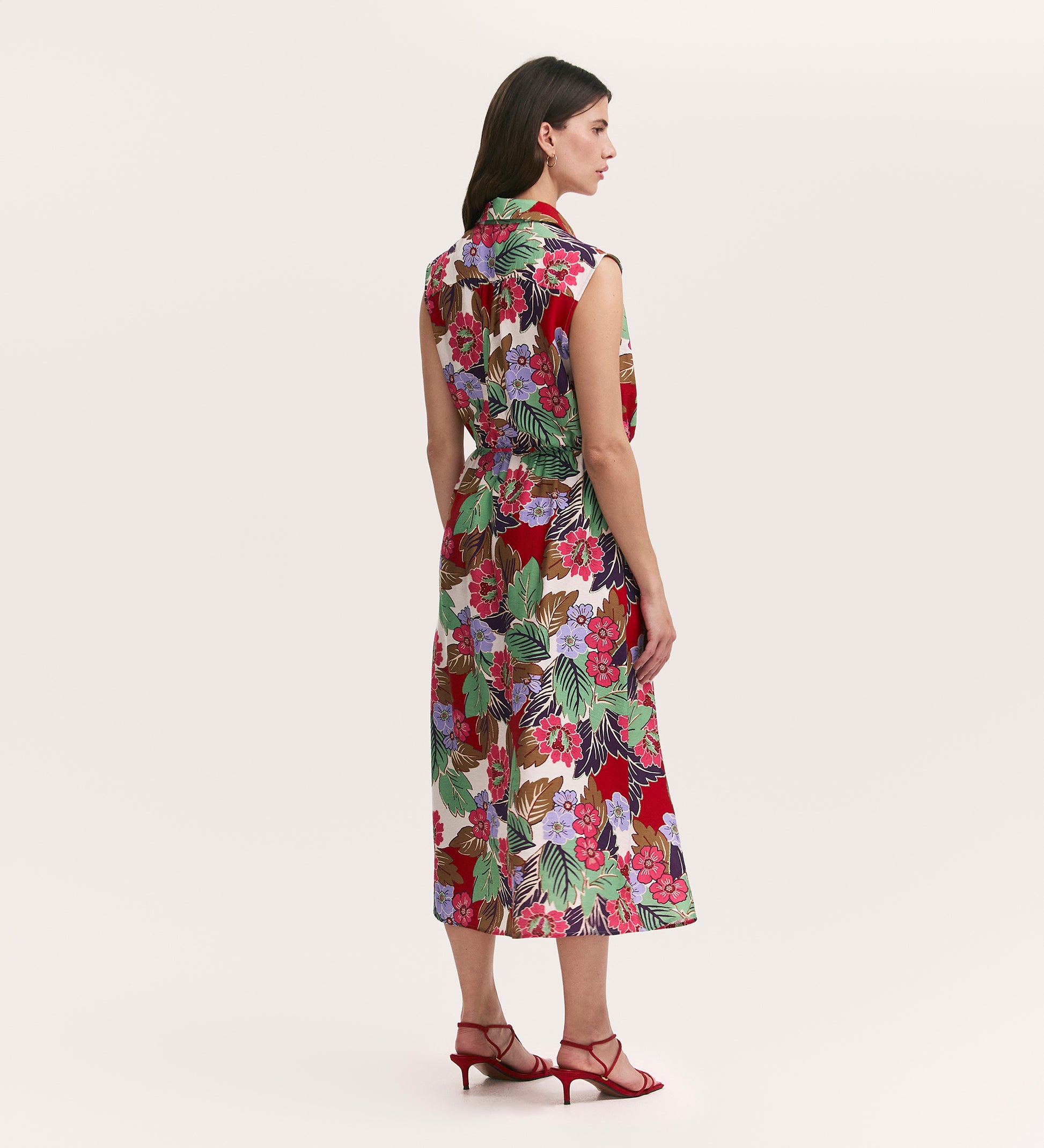A woman with brown hair faces away, wearing the Alida Red Floral Sleeveless Midi Dress with a colorful floral pattern and red strappy heels. The plain, light background highlights the dress’s vibrant design and midi length.