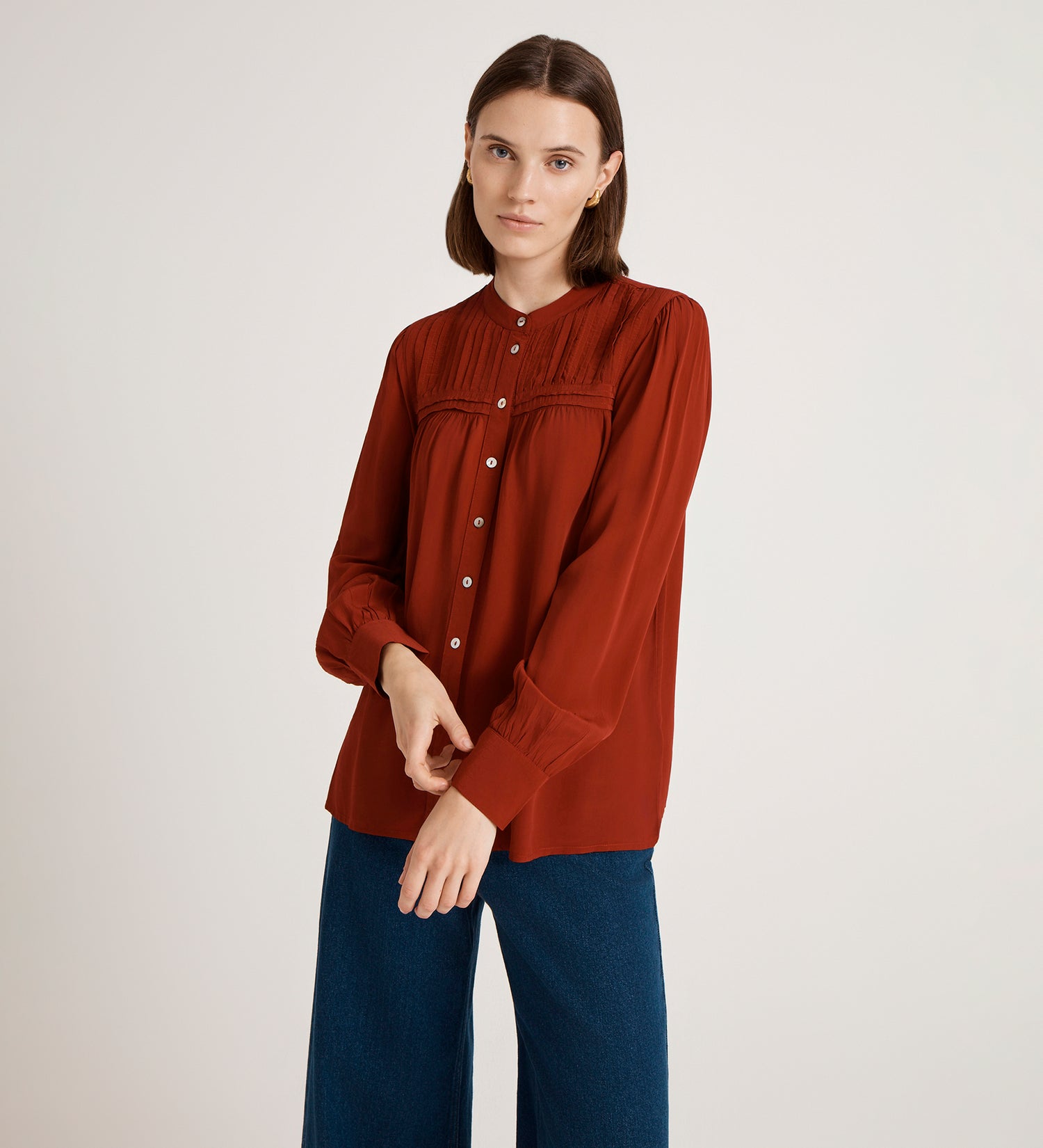 A woman with brown bobbed hair wears the Andrea Red Crepe Blouse with buttons and loose-fitting blue jeans, standing against a plain light background with a neutral expression.