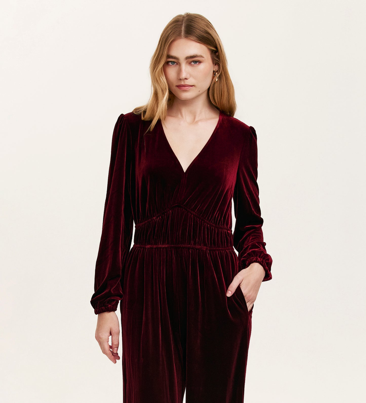 A blonde woman wears the Anna Burgundy Velvet Jumpsuit, featuring wide legs, long sleeves, and a V-neckline. She stands against a plain light background with her left hand in her pocket.