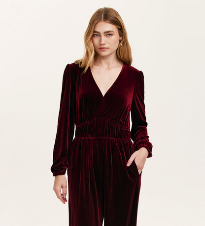 A blonde woman wears the Anna Burgundy Velvet Jumpsuit, featuring wide legs, long sleeves, and a V-neckline. She stands against a plain light background with her left hand in her pocket.