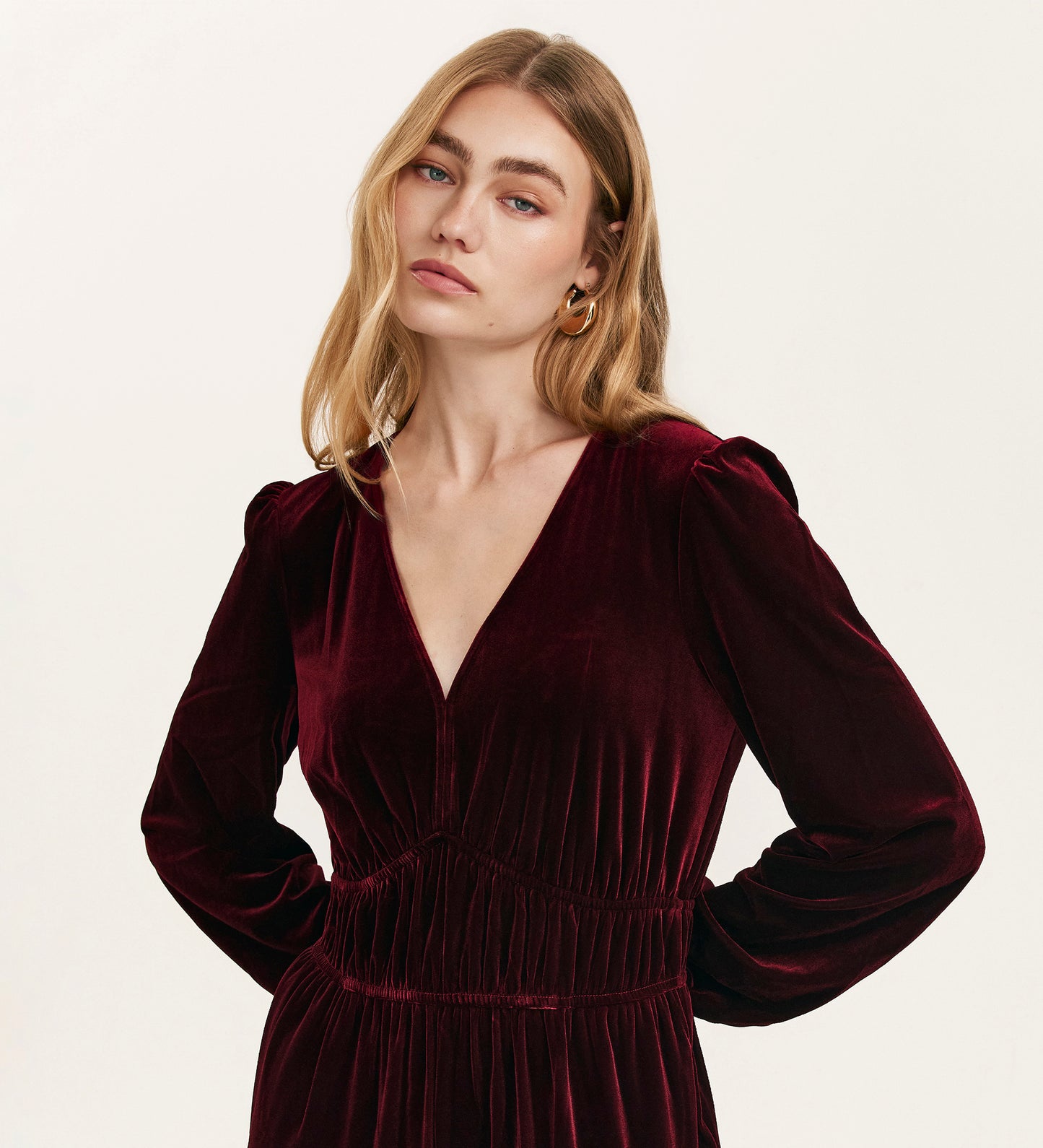 A woman with long blonde hair and gold hoop earrings wears the Anna Burgundy Velvet Jumpsuit, featuring a relaxed fit, long sleeves, and a V-neckline, as she stands thoughtfully against a plain light background.