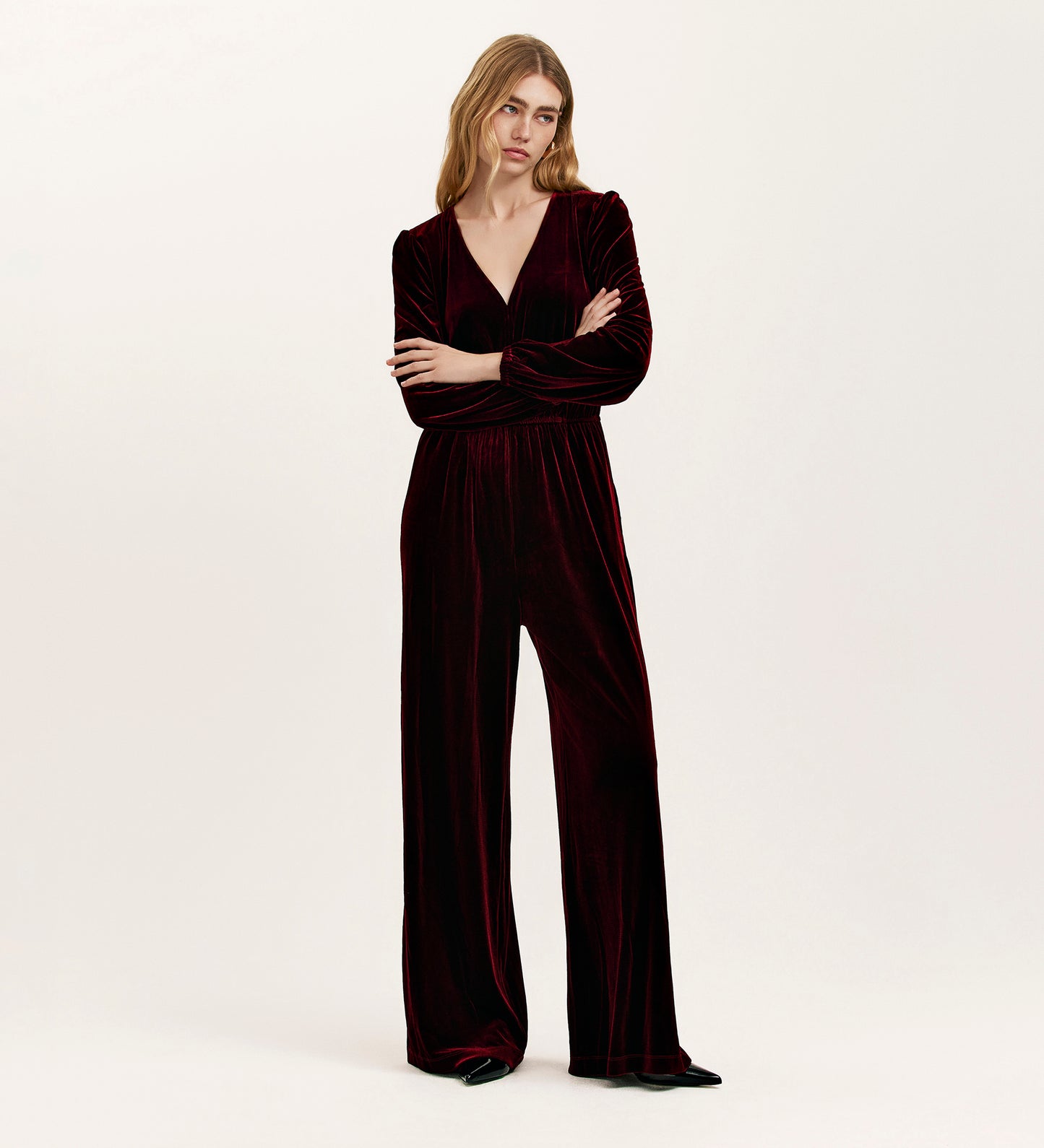 A woman poses against a plain background wearing the Anna Burgundy Velvet Jumpsuit, featuring a relaxed fit, wide legs, and long sleeves. She looks slightly to the side with her arms crossed.