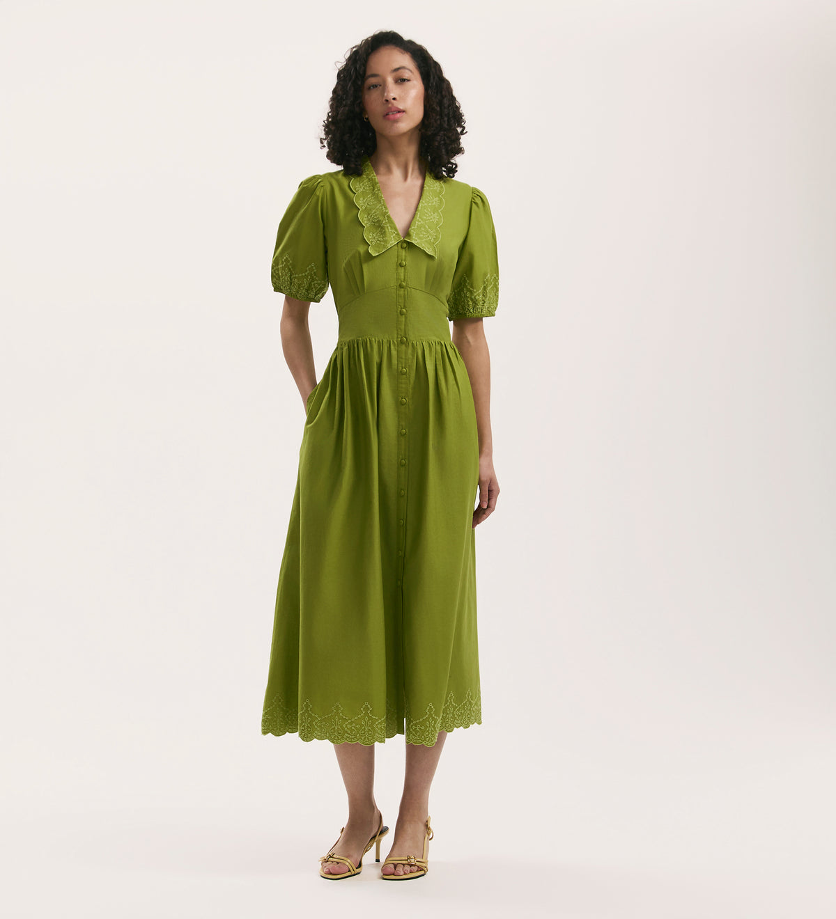 A woman with curly hair stands against a white background, wearing sandals and the Arianna Light Green Embroidered Cotton Midi Dress featuring puffed short sleeves, a lace-trimmed collar, and detailed embroidery on the hem.