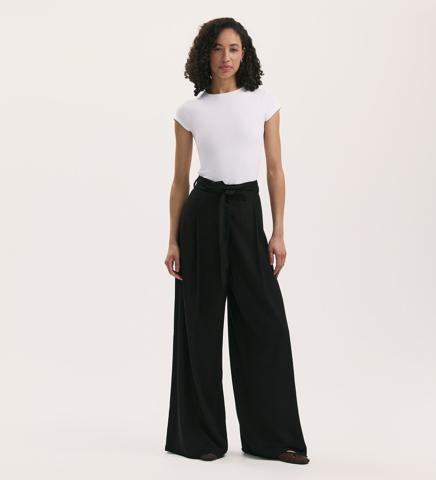 A person with curly hair wears a fitted white short-sleeve shirt and the Athena Black Linen Blend Wide Leg Trousers, standing against a plain white background.