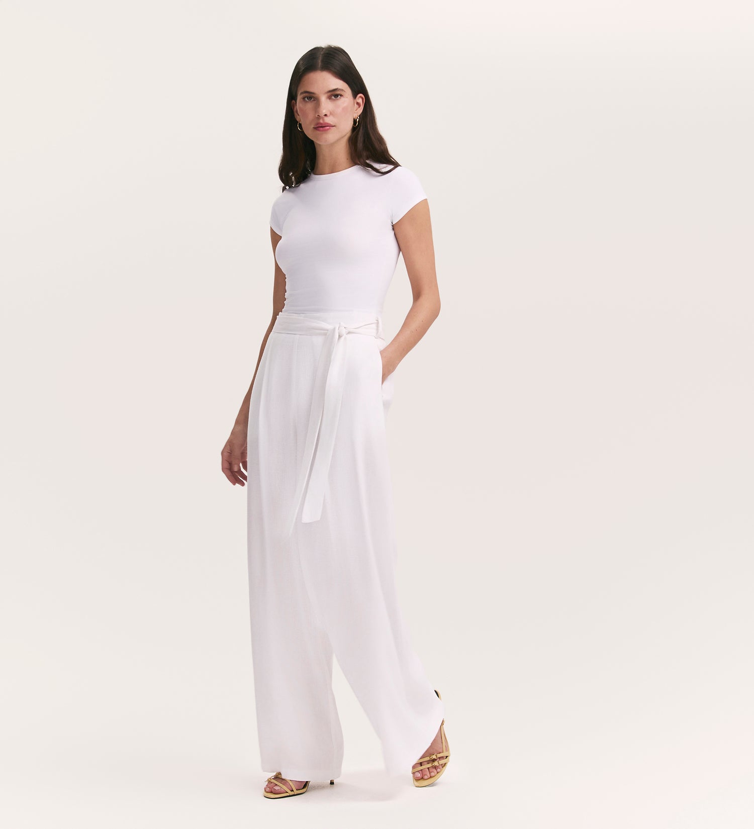 A woman with straight brown hair stands against a light background, wearing Athena White Linen Blend Wide Leg Trousers with a belt and a fitted white short-sleeve top, paired with gold sandals and a neutral expression.