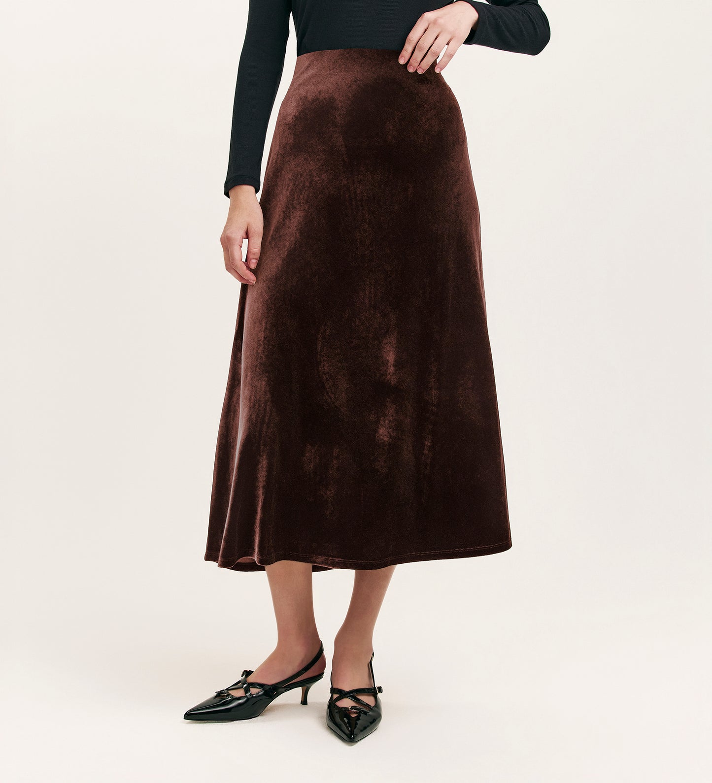 A person wears the Bailey Mocha Jersey Velvet Skirt, featuring a long, dark brown design with an elasticated waistband, styled with a black long-sleeve top and black pointed-toe shoes against a plain light background. Face not visible.