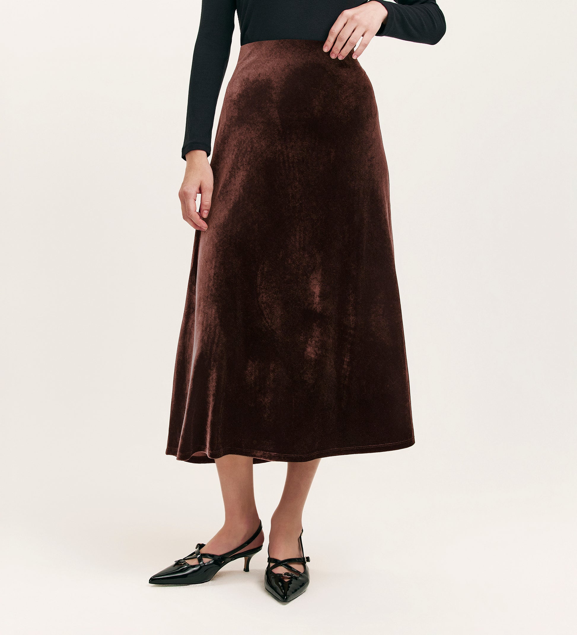 A person wears the Bailey Mocha Jersey Velvet Skirt, featuring a long, dark brown design with an elasticated waistband, styled with a black long-sleeve top and black pointed-toe shoes against a plain light background. Face not visible.