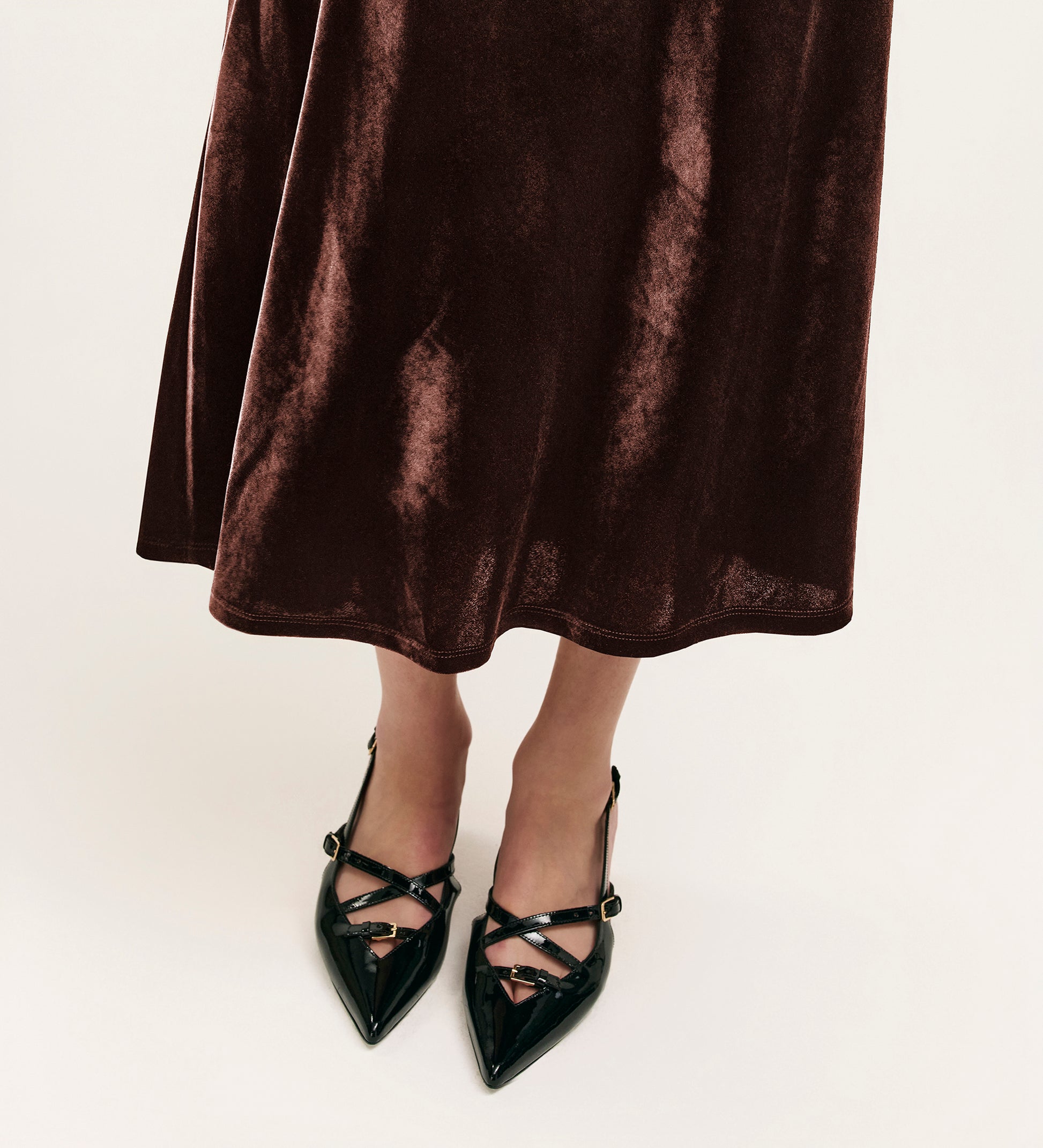 A person wearing the Bailey Mocha Jersey Velvet Skirt and black pointed-toe heels with crisscross straps stands on a light-colored floor.