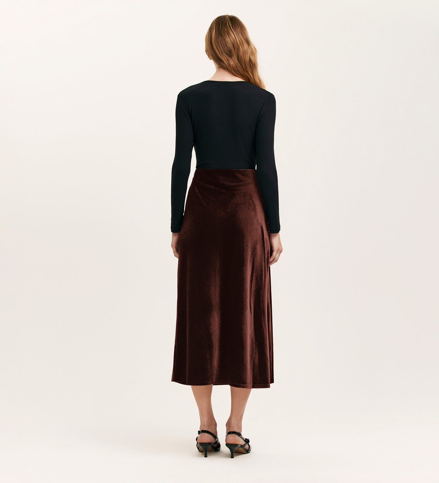 A woman faces away, wearing a long-sleeve black top and the Bailey Mocha Jersey Velvet Skirt—an A-line brown midi with an elasticated waistband—paired with black heeled sandals. The background is plain and light-colored.