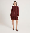 A woman poses in a studio wearing the Beccy Burgundy Crepe Short Dress, a vintage-inspired button-down with long sleeves, ruffled hem, and matching flats against a plain light backdrop.