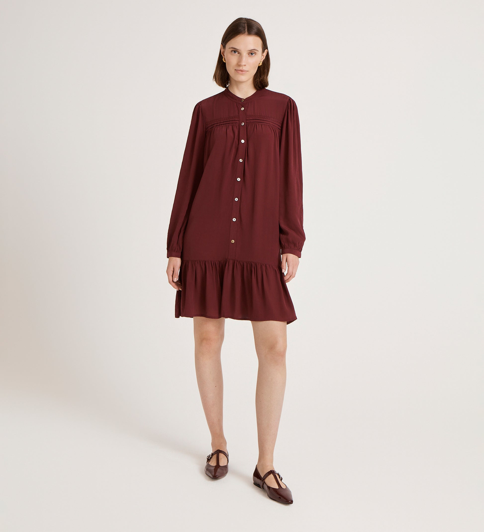 A woman poses in a studio wearing the Beccy Burgundy Crepe Short Dress, a vintage-inspired button-down with long sleeves, ruffled hem, and matching flats against a plain light backdrop.