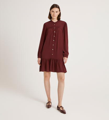 A woman poses in a studio wearing the Beccy Burgundy Crepe Short Dress, a vintage-inspired button-down with long sleeves, ruffled hem, and matching flats against a plain light backdrop.