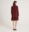 A woman stands facing away, wearing the Beccy Burgundy Crepe Short Dress with long sleeves, a ruffled hem, and brown flats against a plain light background.