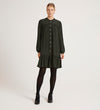 A woman stands against a plain white background, facing forward with her hands by her sides, wearing the Beccy Forest Green Crepe Short Dress—loose-fitting with a mandarin collar, long sleeves, ruffled hem—plus black tights and heeled shoes.