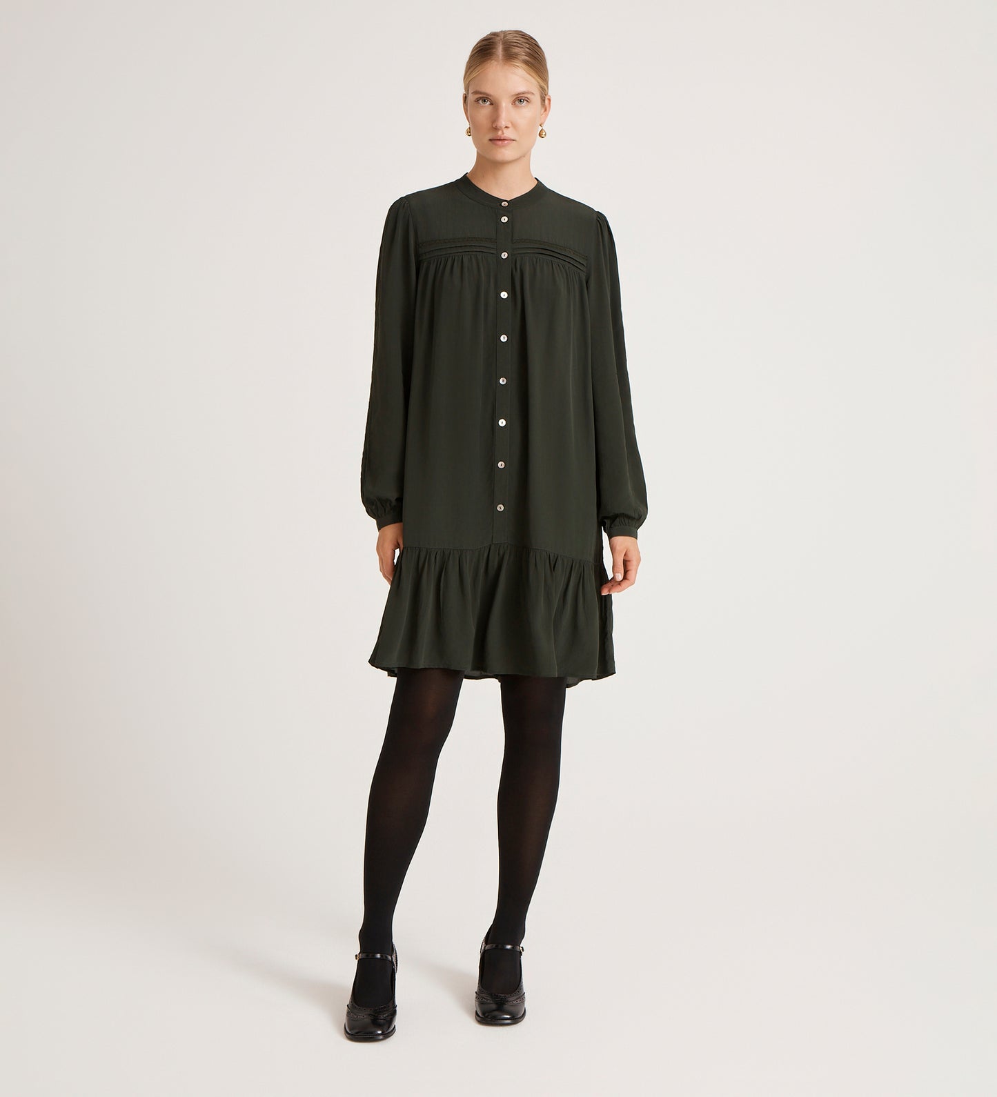 A woman stands against a plain white background, facing forward with her hands by her sides, wearing the Beccy Forest Green Crepe Short Dress—loose-fitting with a mandarin collar, long sleeves, ruffled hem—plus black tights and heeled shoes.