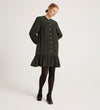 A woman stands in a studio wearing the Beccy Forest Green Crepe Short Dress with black tights and heels. The dress has long sleeves, a mandarin collar, white buttons, and a ruffled hem. She looks down with one foot slightly forward.
