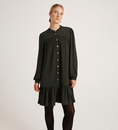 A woman wears the Beccy Forest Green Crepe Short Dress, featuring long sleeves, a loose fit, mandarin collar, button-down front, and ruffled hem, standing against a plain light background.