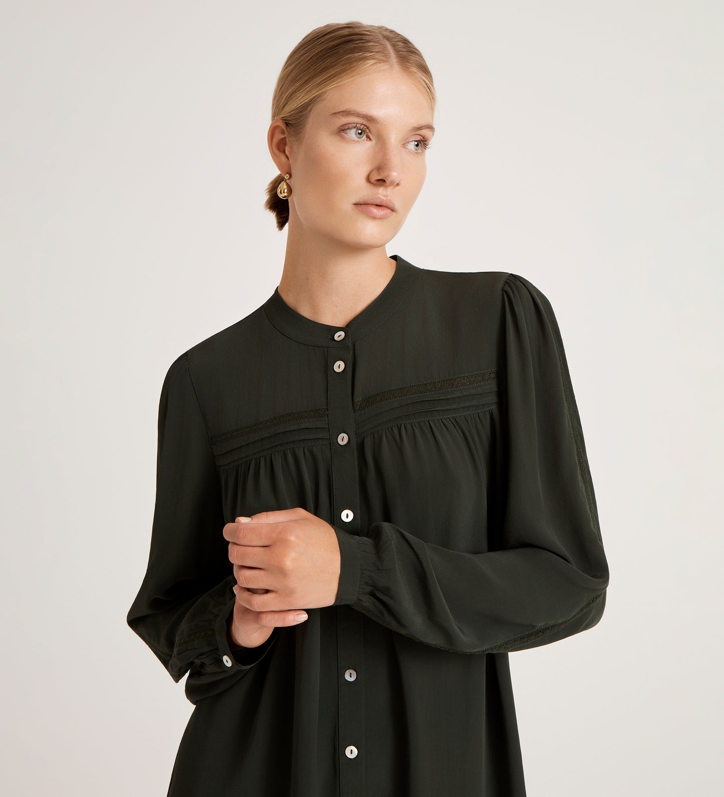 A woman with blonde hair in a low bun wears the Beccy Forest Green Crepe Short Dress and gold earrings, standing against a plain light background and looking to the side with a calm expression.