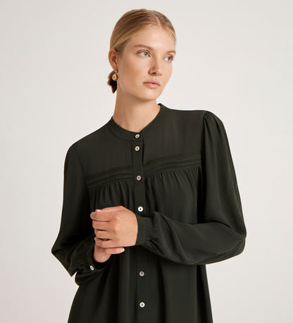 A woman with blonde hair in a low bun wears the Beccy Forest Green Crepe Short Dress and gold earrings, standing against a plain light background and looking to the side with a calm expression.